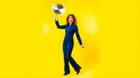 Cheerful young Caucasian woman with curly red hair in blue jumpsuit spinning a silver pan on yellow background, full length.