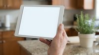 Person holding blank tablet with white screen in modern kitchen interior with wooden cabinets and countertop for app mockup or web design display.