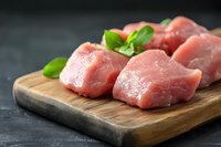 Fresh raw pork tenderloin pieces on wooden cutting board with basil leaves, dark background, close up view for cooking or restaurant menu design.