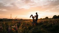 Man lifting woman in wildflower field at golden hour sunset, romantic outdoor engagement or wedding celebration moment in nature.