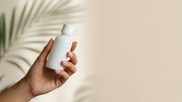 Generative AI image of a hand holding a white cosmetic bottle against a beige background with palm leaf shadows, suitable for skincare, beauty, and wellness product marketing.