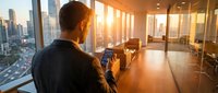 Man in casual dark clothing photographing urban skyline from high-rise office during golden hour sunset, capturing cityscape through smartphone for business or travel content.