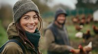 Young Caucasian woman in warm winter clothing smiling at camera while working on organic chicken farm, with coworker and free-range hens in background. Sustainable agriculture.