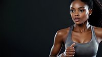 Athletic African American woman in gray sports bra showing thumbs up against dark background, expressing positivity and confidence during workout session.