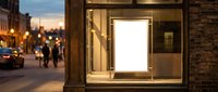 Illuminated blank white poster frame in modern storefront window display at twilight with urban street scene and pedestrians in background.