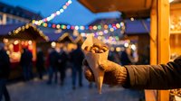 Hands holding warm roasted chestnuts in paper cone at festive Christmas market with colorful lights and wooden stalls creating cozy winter atmosphere.