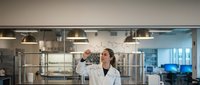 Female scientist in white lab coat examining test tube in modern laboratory with professional equipment and glass partitions for research analysis.