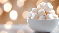 White fluffy marshmallows piled in ceramic bowl against soft bokeh background, creating cozy winter mood for food and beverage photography.