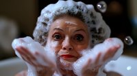Caucasian woman with soap foam on head and hands playfully poses during bath time, expressing joy and relaxation with bubbles floating around her face.