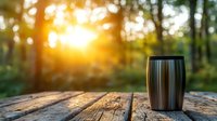 Stainless steel travel mug on rustic wooden table outdoors at sunset with forest bokeh background. Golden sunlight streaming through trees creates cozy atmosphere.