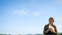 Young woman with blonde hair wearing eyeglasses and olive green jacket holds chicken outdoors under blue sky, suitable for agriculture, farming, and rural lifestyle content.