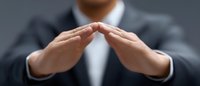 Professional in dark business suit making protective gesture with hands above, demonstrating leadership and security concept against gray background.