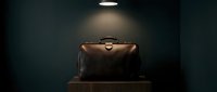 Vintage dark brown leather doctor bag on wooden surface under dramatic spotlight against deep teal wall, evoking mystery and elegance.