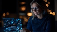 Mature businesswoman in glasses analyzing futuristic holographic car interface projection on digital tablet screen in dark office with bokeh lights.