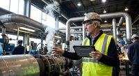Senior industrial engineer in safety vest using tablet computer while inspecting steam pipes and machinery in modern manufacturing facility.