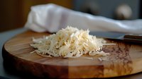 Freshly grated white cheese piled on rustic wooden cutting board with kitchen knife and white cloth in soft natural lighting, food preparation concept.