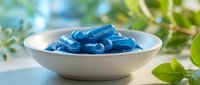 Blue gel capsules in white ceramic bowl on natural background with green leaves for health and wellness supplement photography.