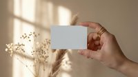 Female hand holding blank white business card mockup with golden rings and dried flowers in soft natural sunlight for branding design templates.