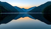 Tranquil mountain lake at dawn with perfect mirror reflection of silhouetted peaks and golden sky in calm waters, creating symmetrical landscape composition.