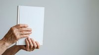 Mature man with wrinkled hands holding blank white book or magazine against neutral background, suitable for publishing, education, and marketing materials.