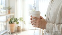 Woman in beige sweater holding white coffee cup in bright modern home interior with plants and natural light streaming through windows.