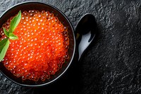 Fresh red caviar in black ceramic bowl garnished with basil leaf on dark stone background, luxury seafood delicacy with copy space.