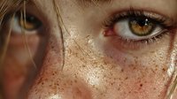 Close-up of amber eye with natural freckles across fair skin, strands of blonde hair falling across face, showing authentic beauty detail.