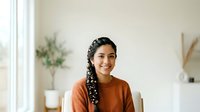 Young woman with long dark braided hair and medium brown skin tone wearing a rust-colored sweater, smiling at camera in bright minimalist interior with plants and natural light.