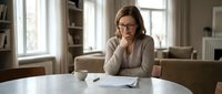 Middle-aged Caucasian businesswoman with glasses reading documents at home office desk, thoughtful expression, natural lighting from window.