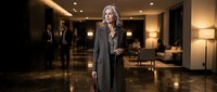 Professional blonde businesswoman in elegant coat standing confidently in luxury hotel lobby with marble floors and warm lighting for corporate concepts.