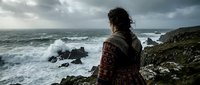 Woman in traditional patterned clothing contemplating stormy ocean waves from rocky coastal cliff under dramatic cloudy sky for mindfulness and travel concepts.