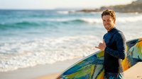 Man in wetsuit holding colorful surfboard on sandy beach with ocean waves and coastal cliffs in background. Surfer ready for water sports activity.