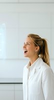 Young woman with blonde hair in ponytail, wearing white button-up shirt, smiling with mouth open in professional office setting near whiteboard, suitable for business and corporate communications.