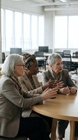 Three mature women of diverse ethnicities engaged in discussion at wooden table in modern office space with large windows and contemporary furniture.