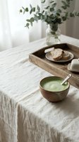 Minimalist breakfast table setting with green matcha soup in wooden bowl, sliced bread on ceramic plate, fresh eucalyptus branches in glass vase, and linen tablecloth for home decor and lifestyle content.