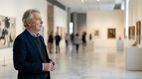 Senior man with gray hair and light skin tone, wearing dark blazer, viewing artwork in contemporary gallery space with polished floors and blurred visitors in background.