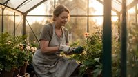 Middle-aged Caucasian woman gardener tending plants in sunny greenhouse wearing apron and gloves for organic farming and horticulture activities.