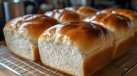 Fresh baked golden milk bread loaves cooling on wire rack, showing soft texture and glossy brown crust, ready for slicing and serving.
