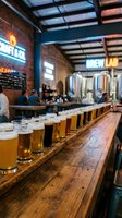 Craft beer tasting flight lined up on wooden bar counter in modern brewery taproom with neon signage and industrial ceiling design.