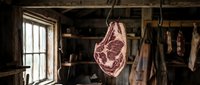 Raw beef cuts hanging in rustic butcher shop with natural light from window, meat curing and processing facility for culinary and food industry use.