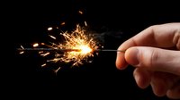 Bright burning sparkler with glowing sparks flying against dark background, hand holding lit firework creating festive light trails and smoke effects.