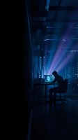 Silhouette of programmer working late at night in dark office with glowing computer screen and neon blue lighting creating atmospheric workspace environment.