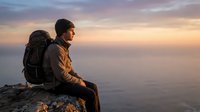 Young male hiker with backpack sitting on mountain peak at sunset, contemplating vast landscape below during golden hour adventure and travel journey.