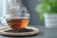 Hot steaming black tea in transparent glass cup on wooden plate with scattered dry tea leaves, blurred green plant background, moody atmosphere.