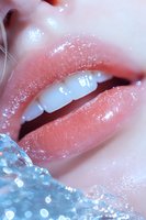 Close-up of glossy coral lips with water droplets and sparkling effects against blue crystal ice formation, showing white teeth in sensual beauty composition.
