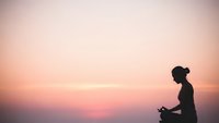 Young woman meditating in lotus pose during sunset with peaceful pink sky background for wellness and mindfulness practices.