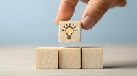 Hand placing wooden cube with light bulb icon on stack of blank blocks against blue background, symbolizing creative idea or innovation concept.