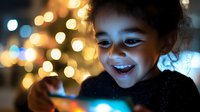 Young mixed-race girl with curly hair smiling while using smartphone with warm bokeh lights creating magical atmosphere for technology and childhood themes.