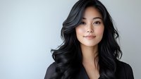 Young Asian woman with flowing black wavy hair and natural makeup wearing black blazer, smiling confidently against light gray background. Professional headshot style portrait.