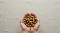 Hands holding handful of almonds against neutral beige background. Suitable for nutrition, healthy eating, snack food, and wellness content.